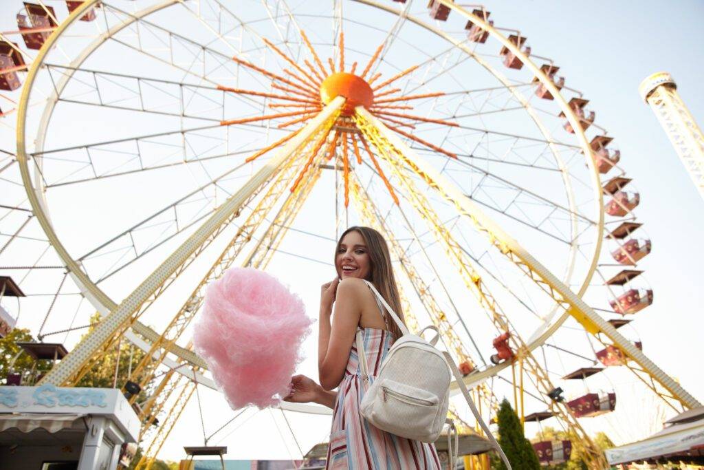 outdoor-shot-happy-young-brunette-lady-with-long-hair-wearing-romantic-dress-white-backpack-standing-ferris-wheel-summer-warm-day-holding-cotton-candy-smiling-widely